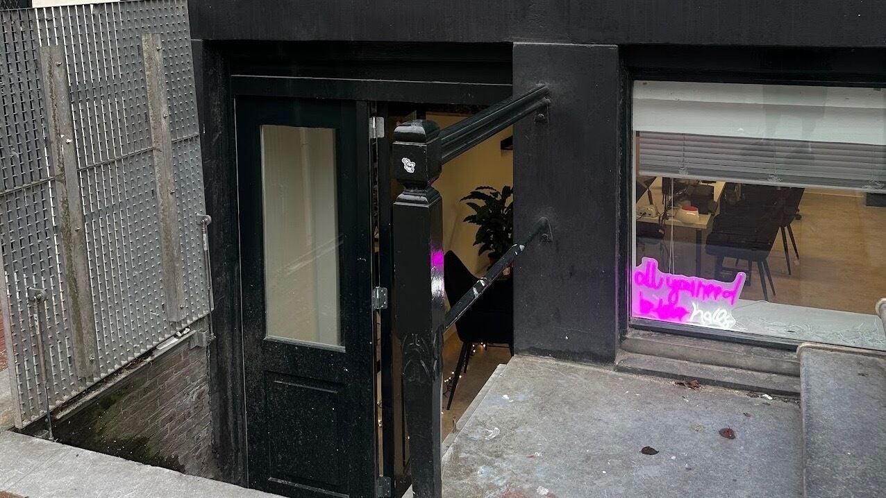 De ingang van Juss Nail: een zwarte deur in het souterrain, met een roze neonbord in het raam